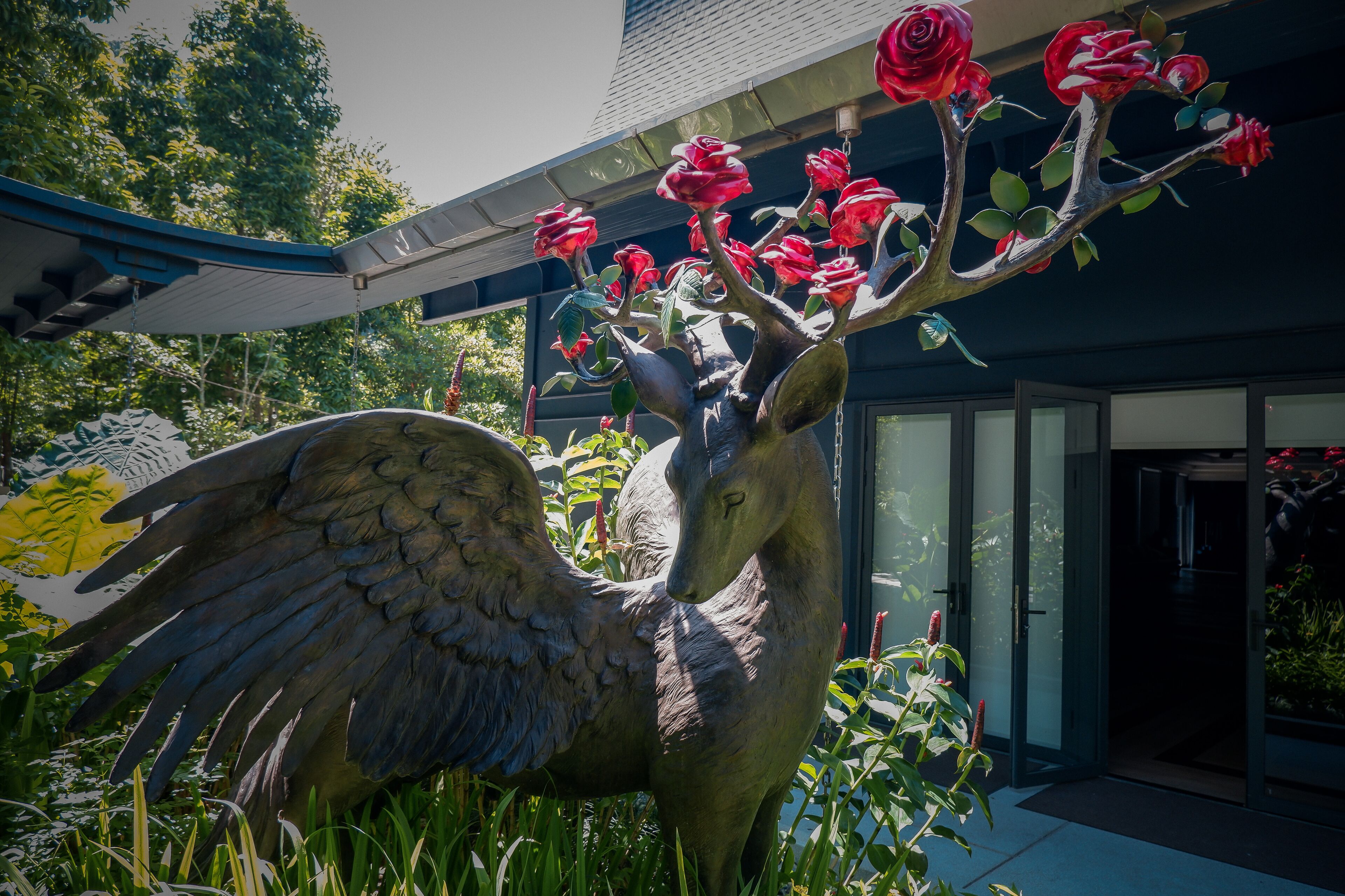 A sculpture of a deer with large wings and red flowers on its antlers, set in a garden with a building in the background.