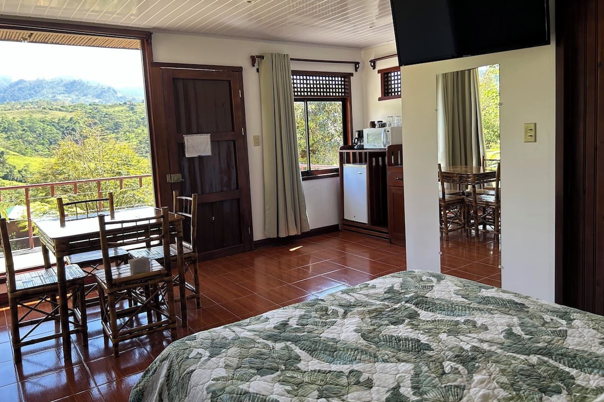 Une chambre à coucher avec un lit, un bureau et une chaise. Il y a une fenêtre donnant sur un paysage verdoyant et une porte.