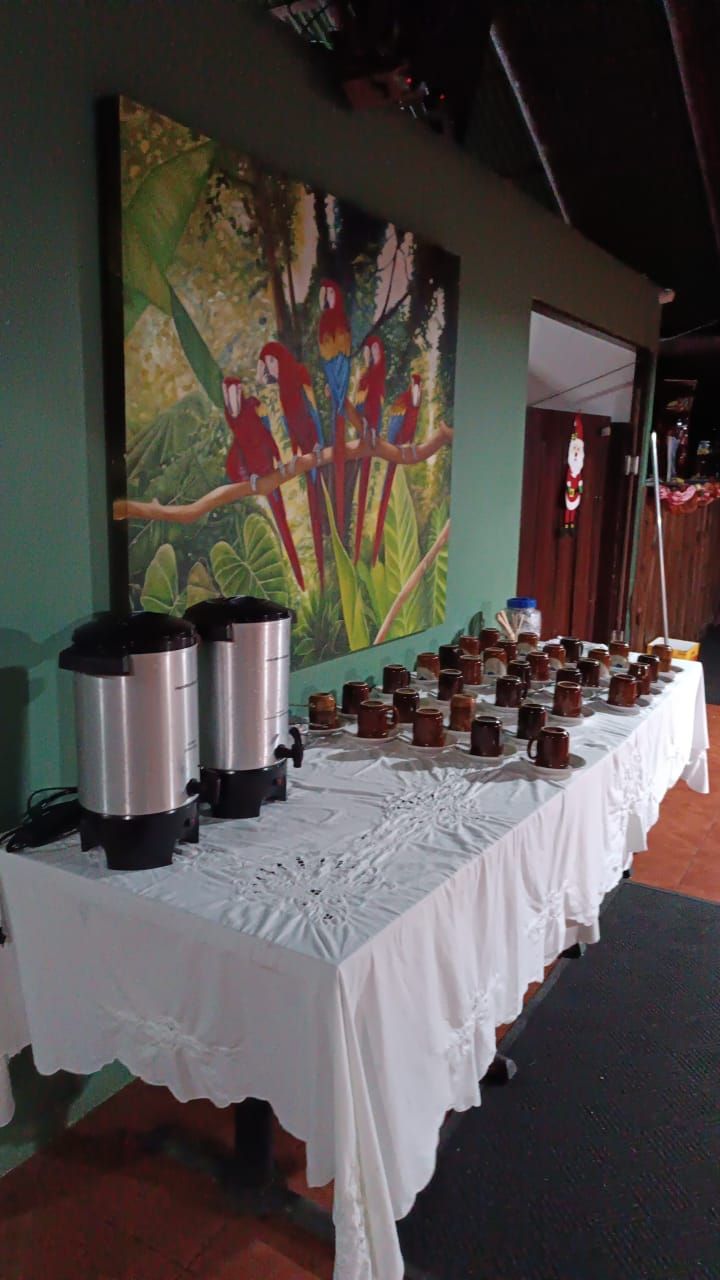 A buffet table with coffee urns and mugs, a large painting of birds on the wall, and a door in the background.