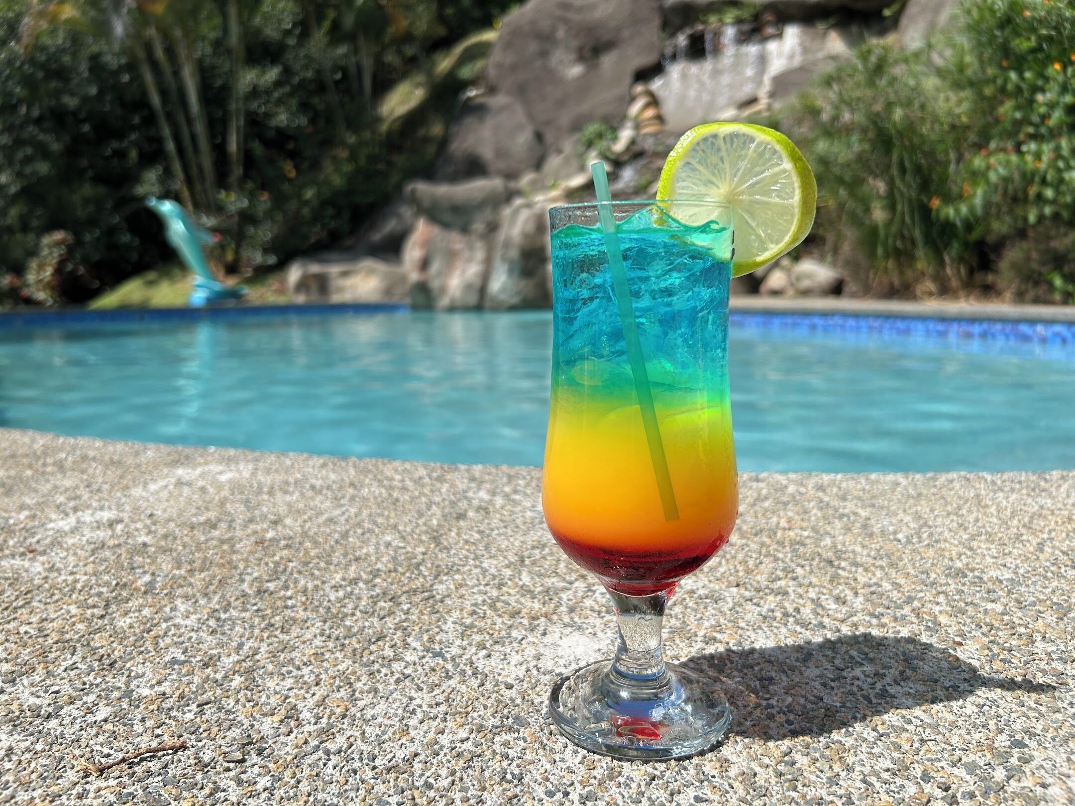A glass of colorful drink with a lime slice, placed on a textured surface near a pool.