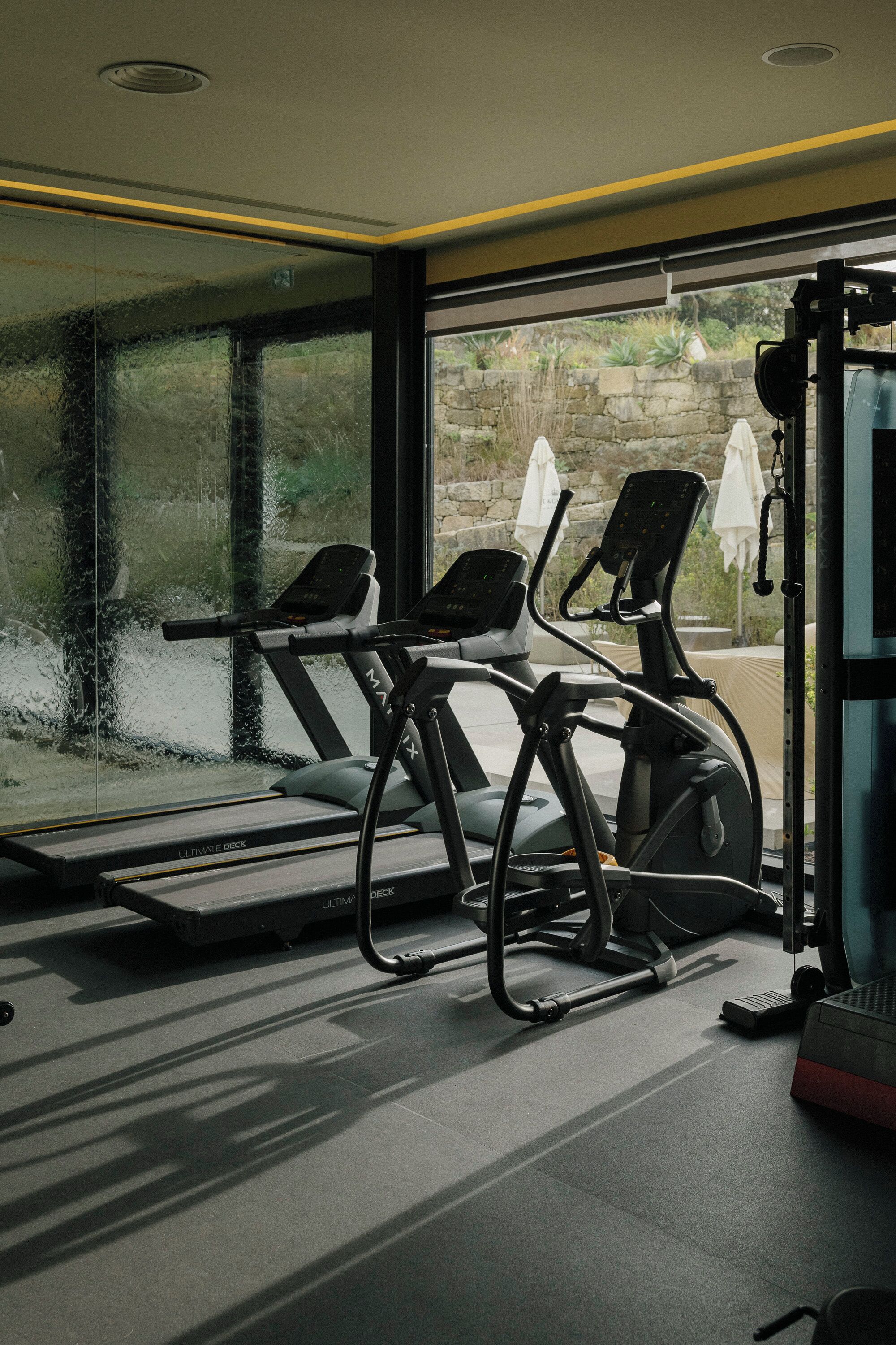 Une salle de sport équipée de tapis de course et d’appareils d’exercice, de grandes fenêtres donnant sur un espace verdoyant et un mur en pierre à l’extérieur.