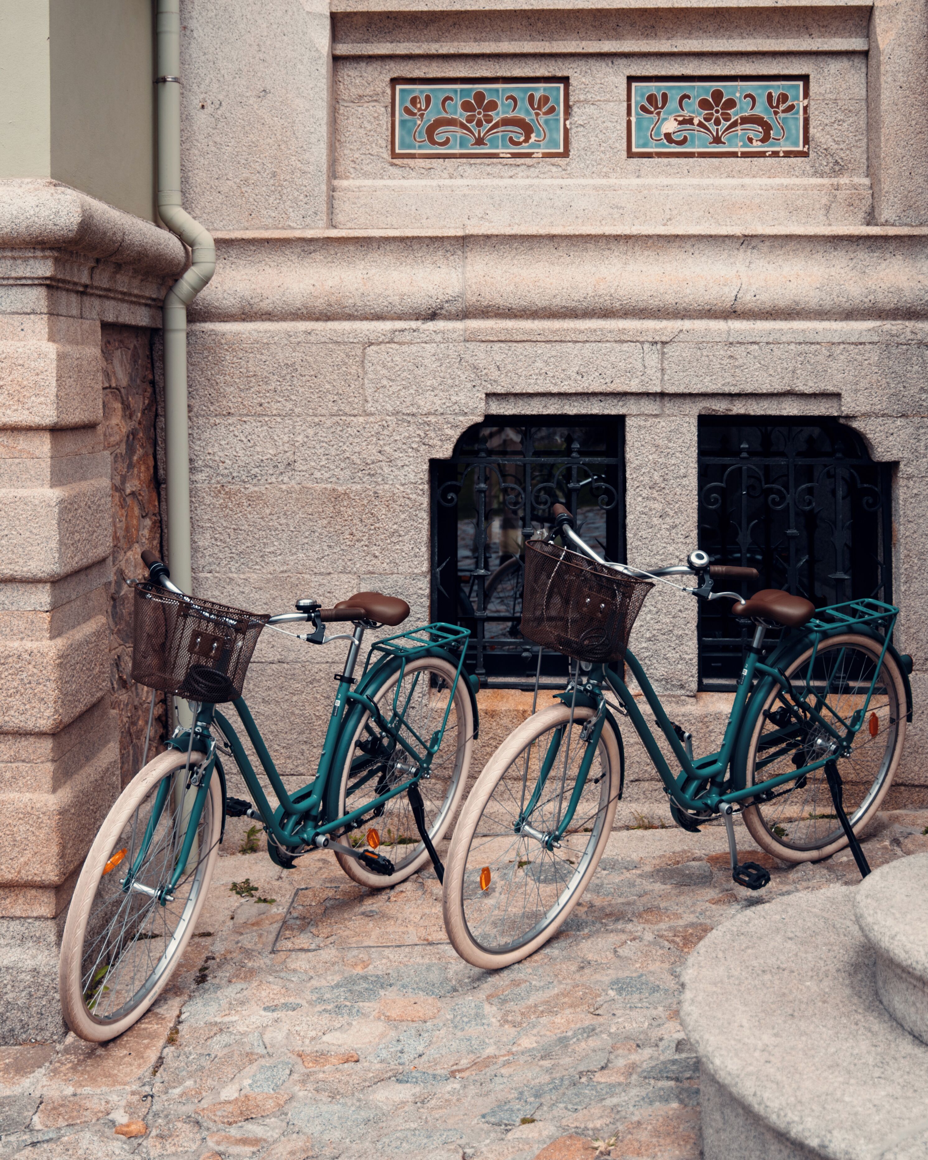 Twee fietsen geparkeerd tegen een stenen muur met een decoratief tegelschilderij boven een zwart ijzeren hek.