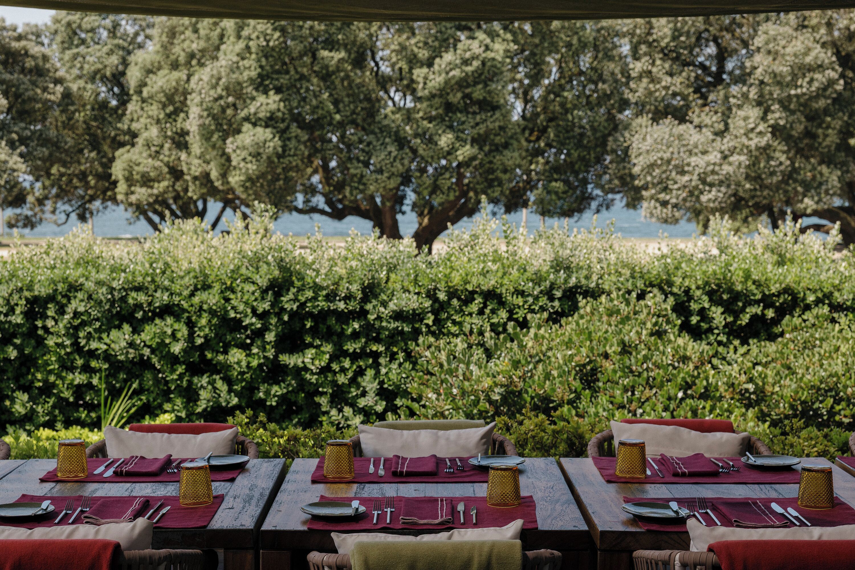 An outdoor dining area with tables set for two, each with a red tablecloth, white plates, and cutlery.