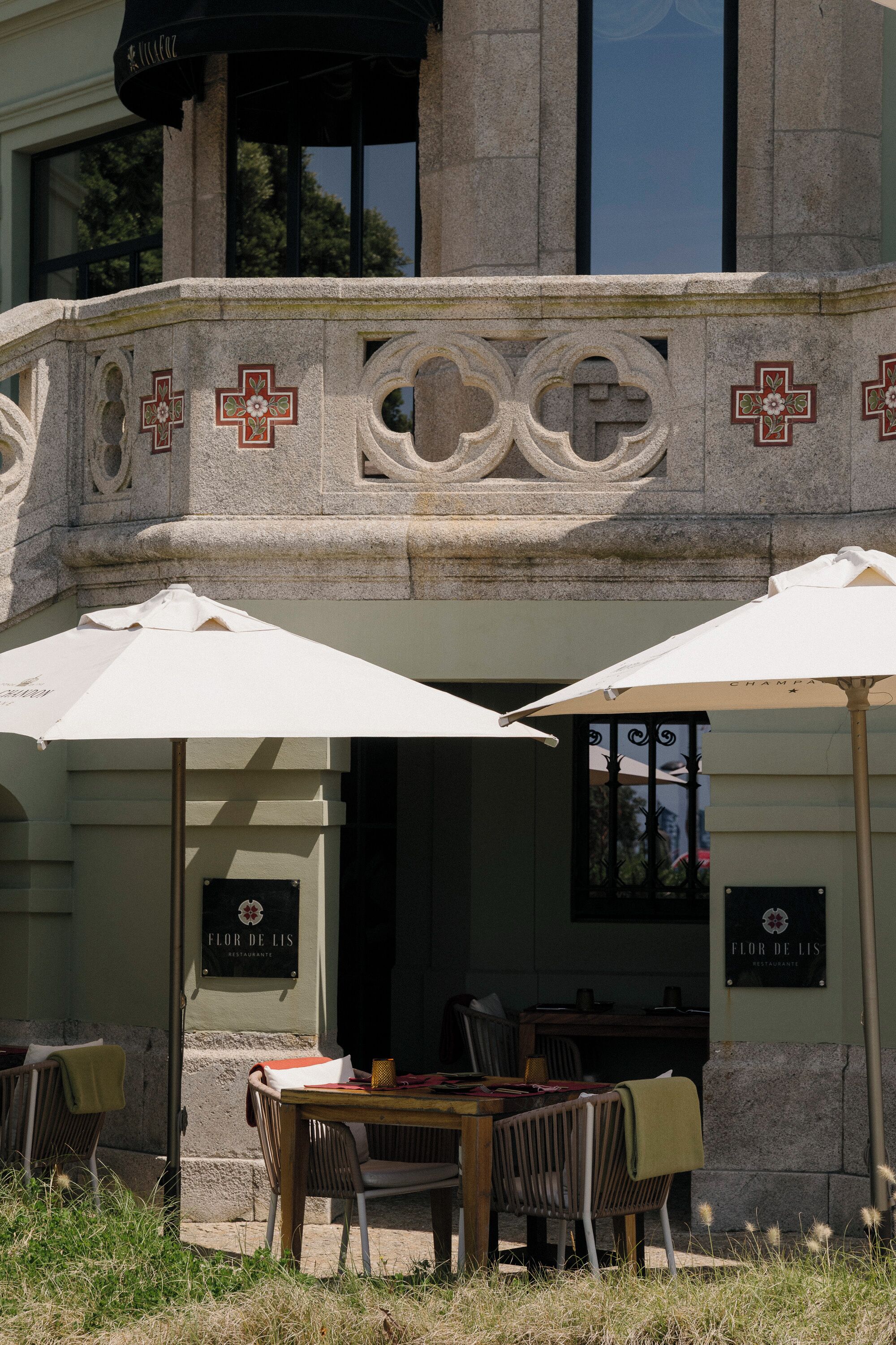 Un área de estar al aire libre con mesas y sillas, sombrillas, y fachada de edificio con detalles de piedra y letrero que dice "Flor de Lis Restaurant".