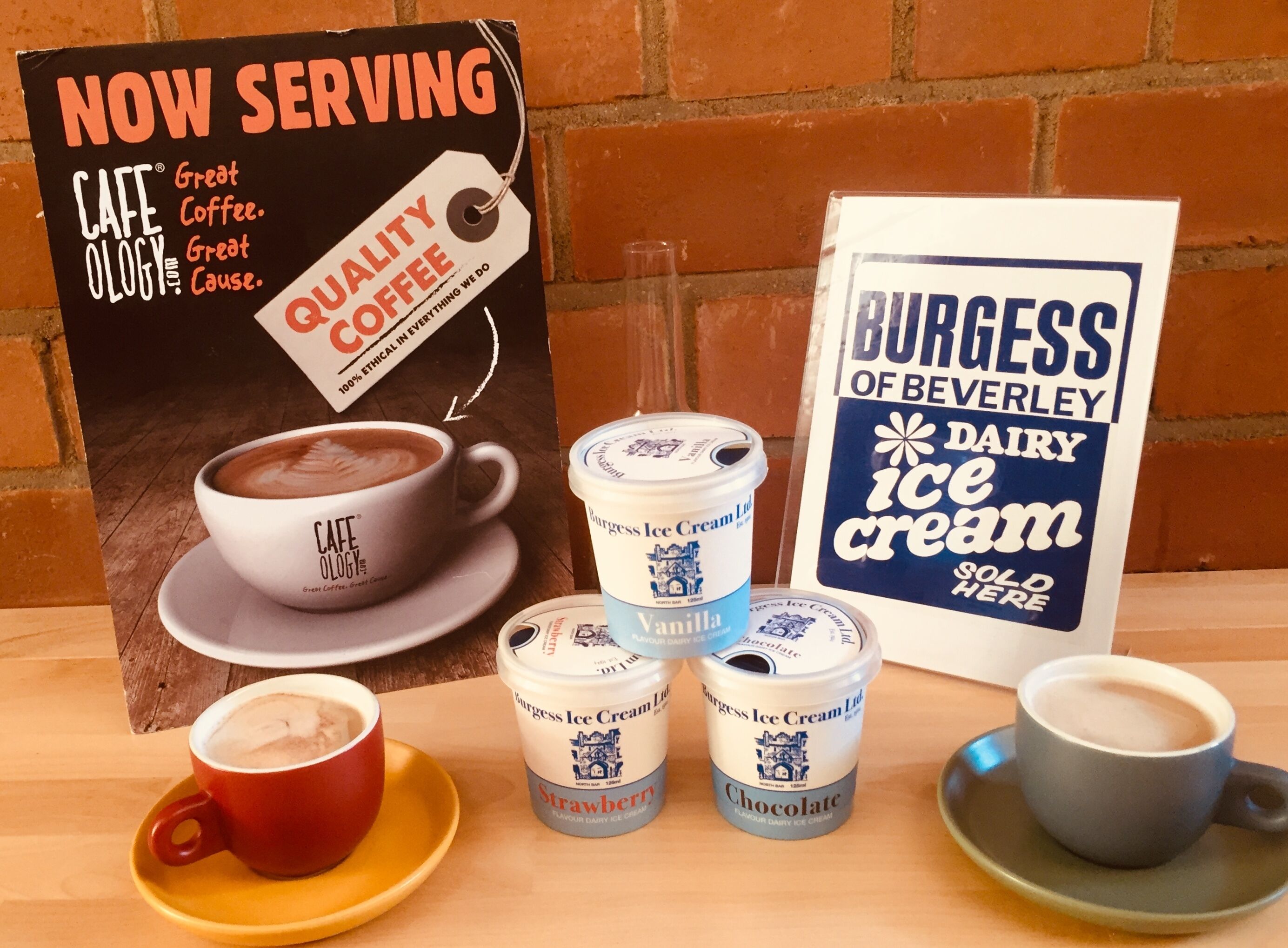 A table with a coffee cup, a sign for Burgess of Beverly Dairy Ice Cream, and three ice cream containers labeled vanilla, strawberry, and chocolate.