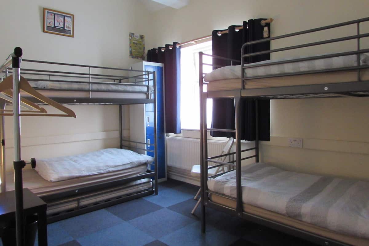 Une chambre de dortoir avec des lits superposés, un radiateur, une fenêtre avec des rideaux et un tableau encadré au mur.
