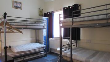 A dormitory room with bunk beds, a radiator, a window with curtains, and a framed picture on the wall.