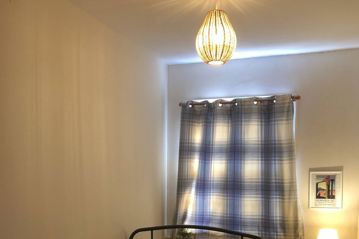 Une chambre à coucher avec un lit, une table de chevet avec une lampe, une fenêtre avec un rideau à carreaux et un radiateur.