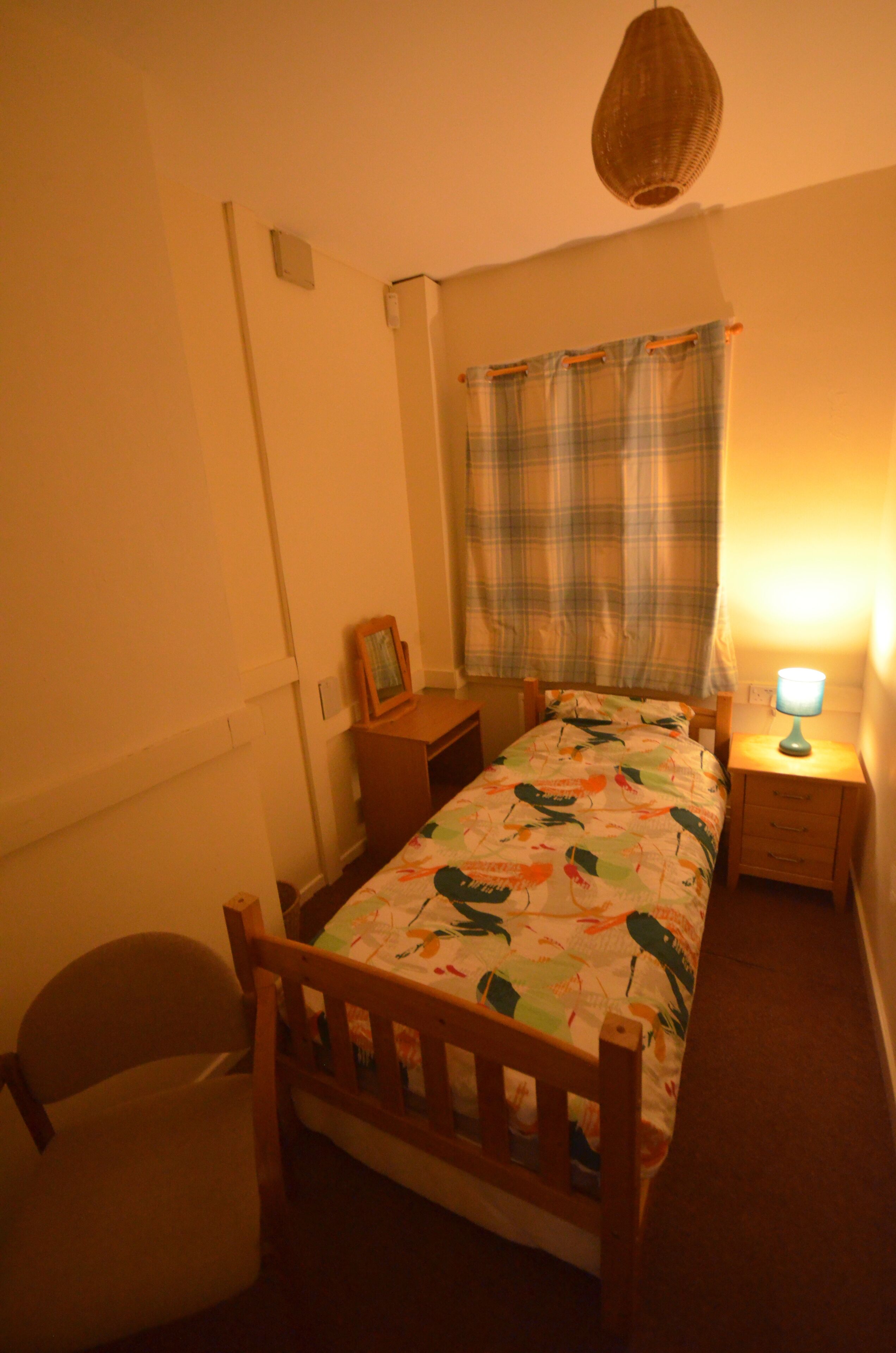 A small, cozy bedroom with a single bed, a nightstand, a chair, and a hanging light fixture.