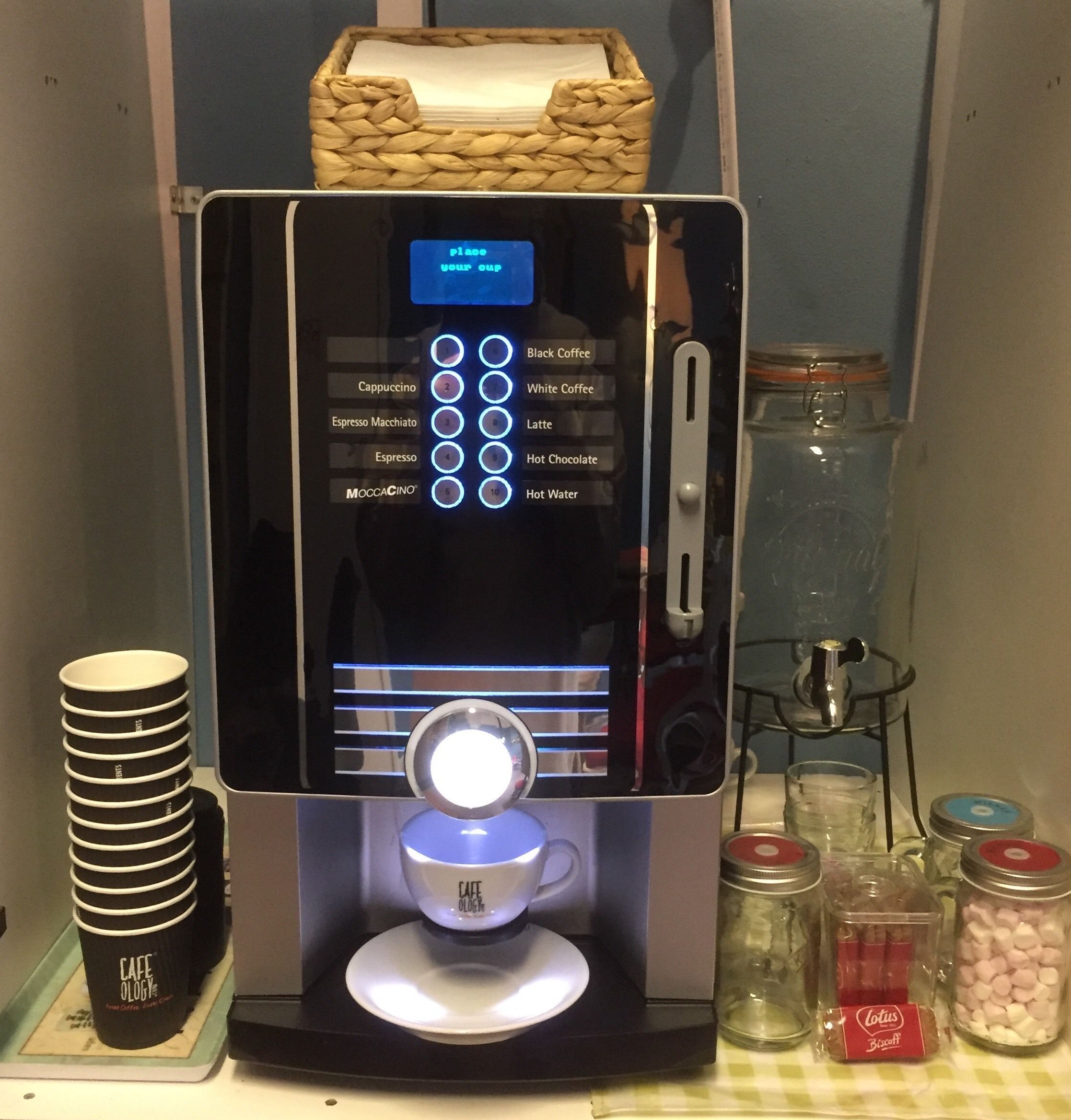 A coffee machine with a digital interface for selecting drinks, a stack of disposable cups, and various condiments.