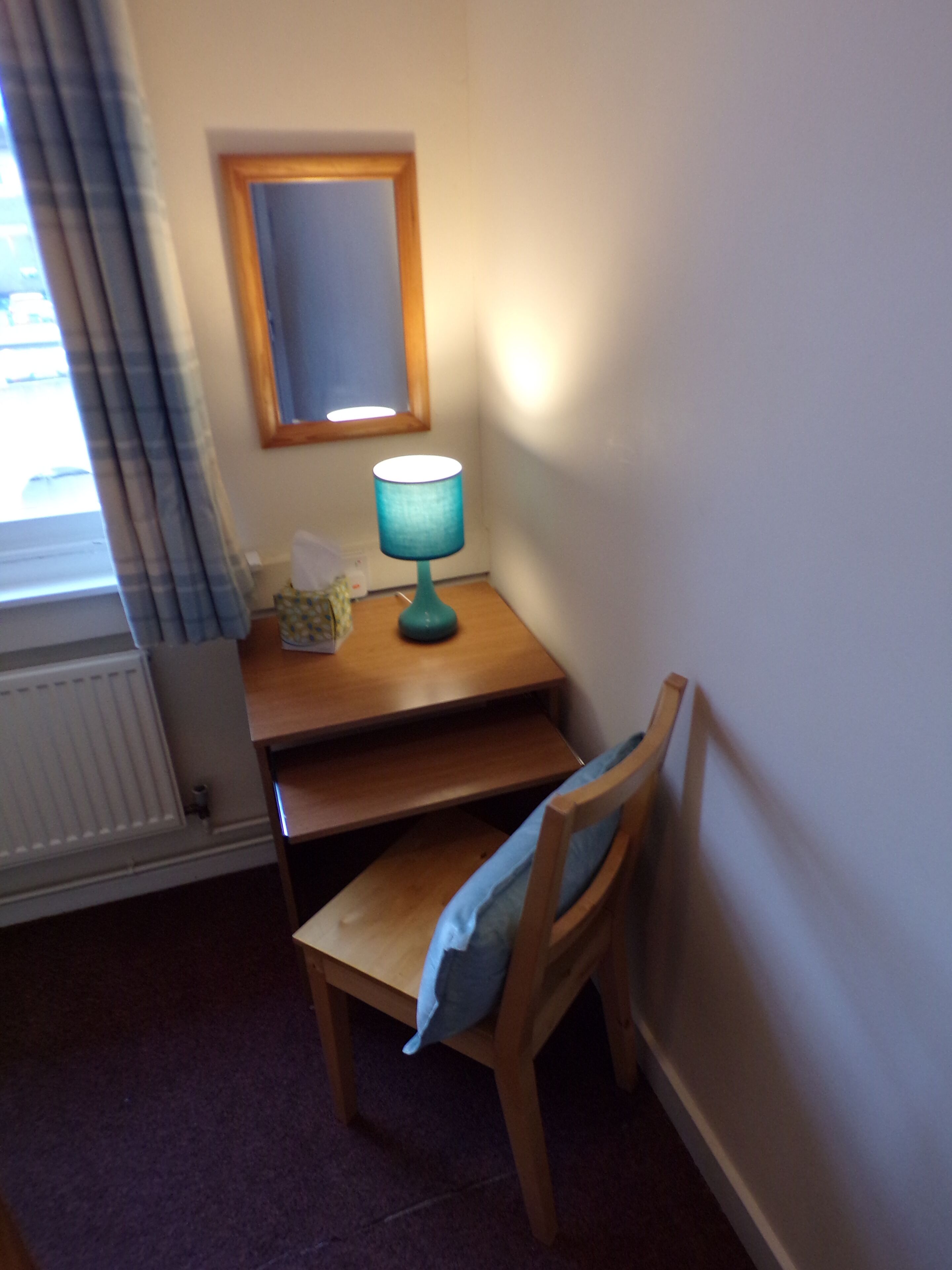 Una habitación pequeña con un escritorio de madera, una silla, una lámpara, un espejo y un radiador.