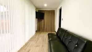 A modern living room with a black leather sofa, a flat-screen TV mounted on the wall, and a large window with sheer curtains. - Pura Natura Lodge (Quepos)