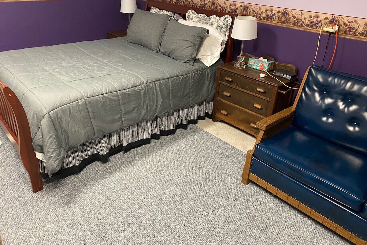 Um quarto com cama, mesa de cabeceira, cômoda e uma cadeira azul.