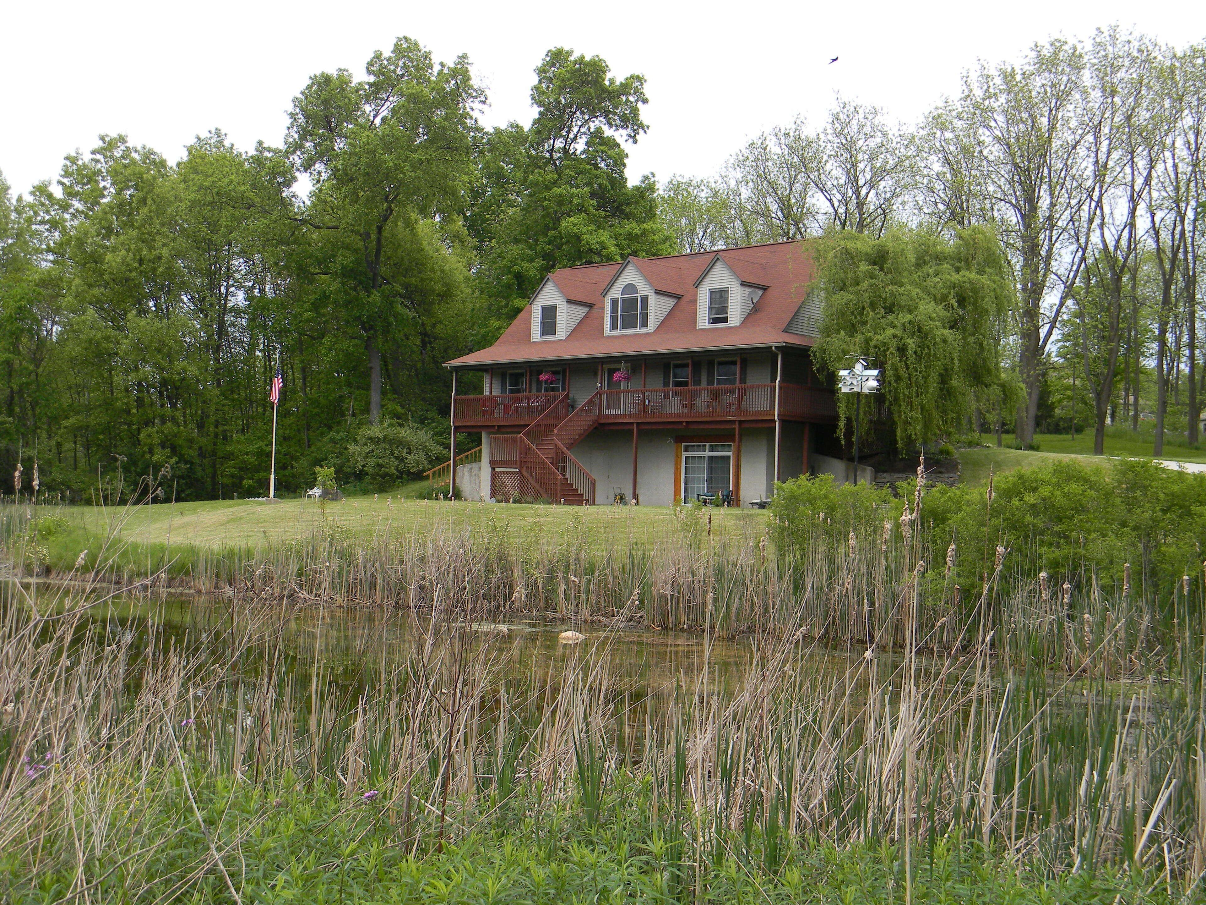 A two-story house with a balcony, surrounded by a pond and trees.