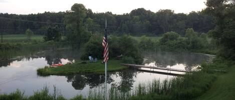 A flagpole with an American flag in a grassy area near a body of water, with a dock and chairs in the background.