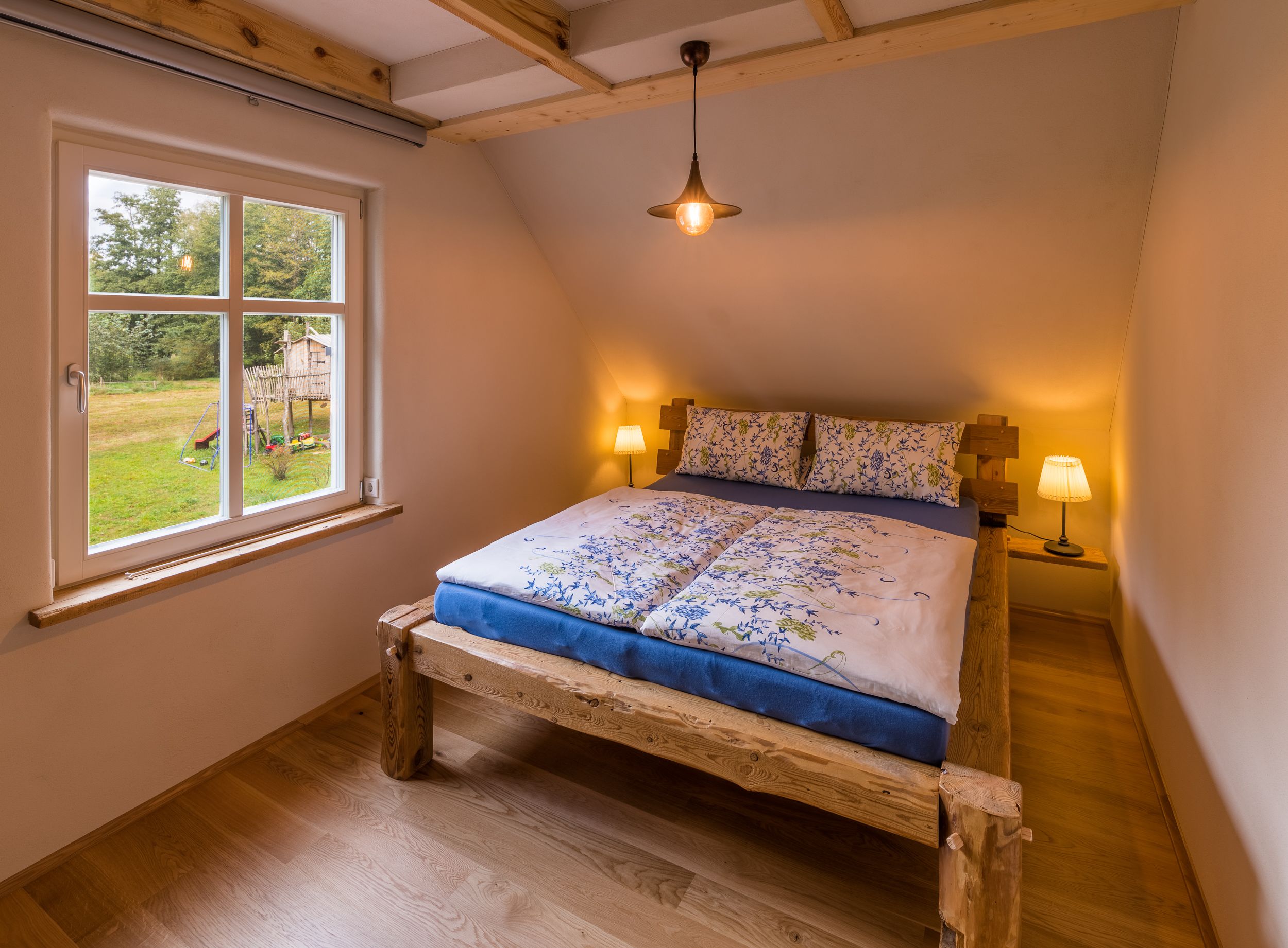 A bedroom with a wooden bed frame, a window with a view of the outdoors, and two bedside lamps.