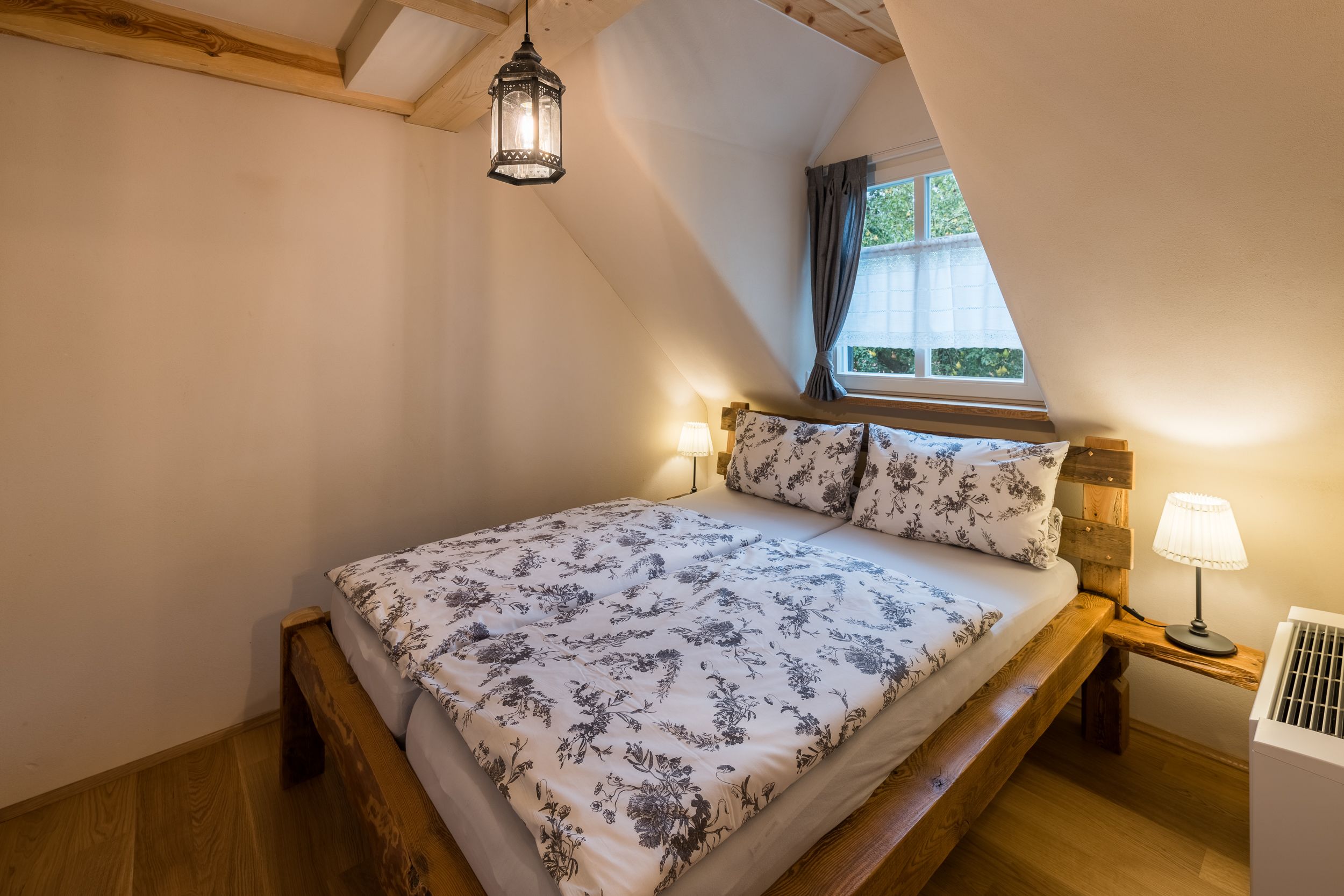 A bedroom with a wooden bed, floral bedding, a nightstand with a lamp, and a window with curtains.