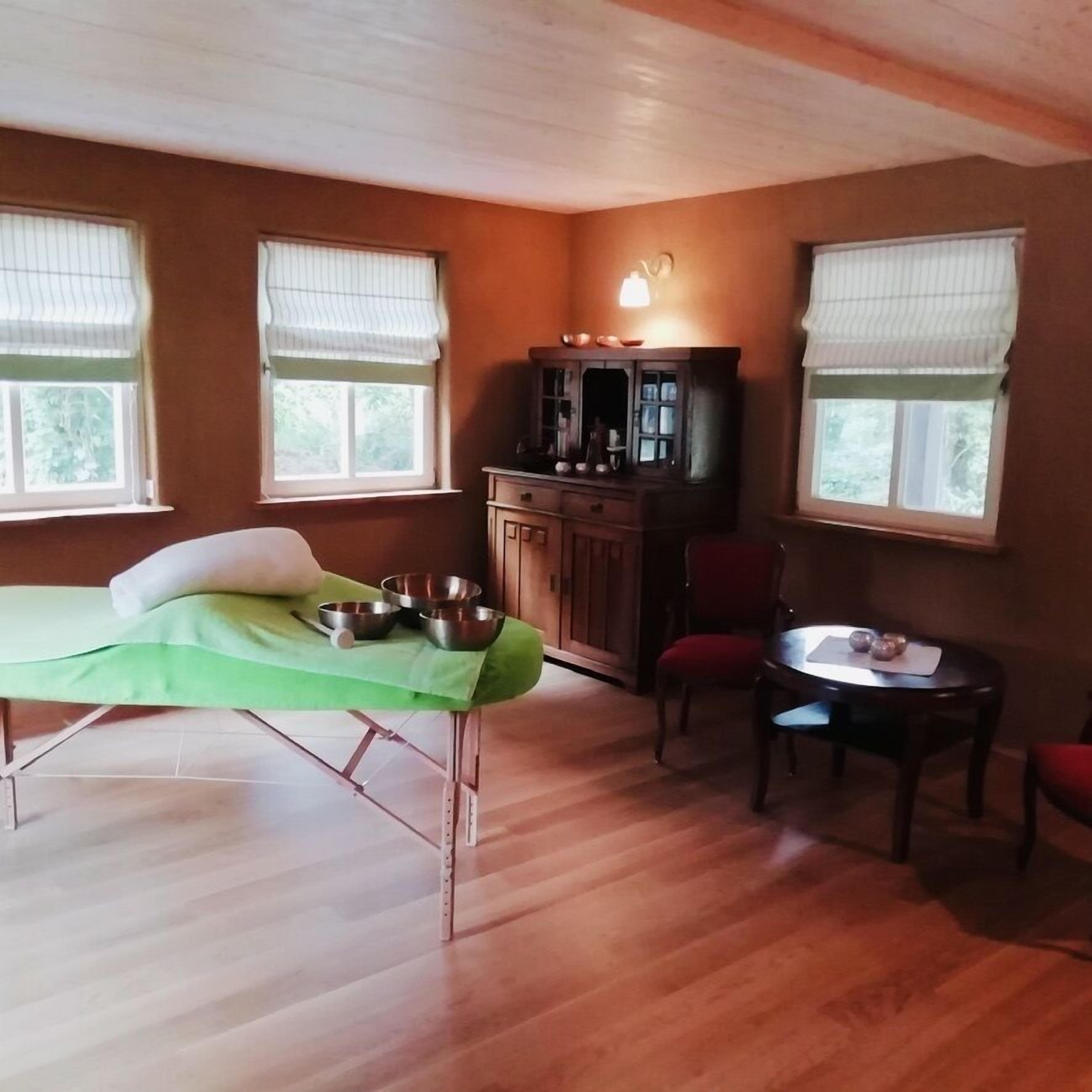 A room with a wooden floor, a green bed, a small table with chairs, and a cabinet with dishes.