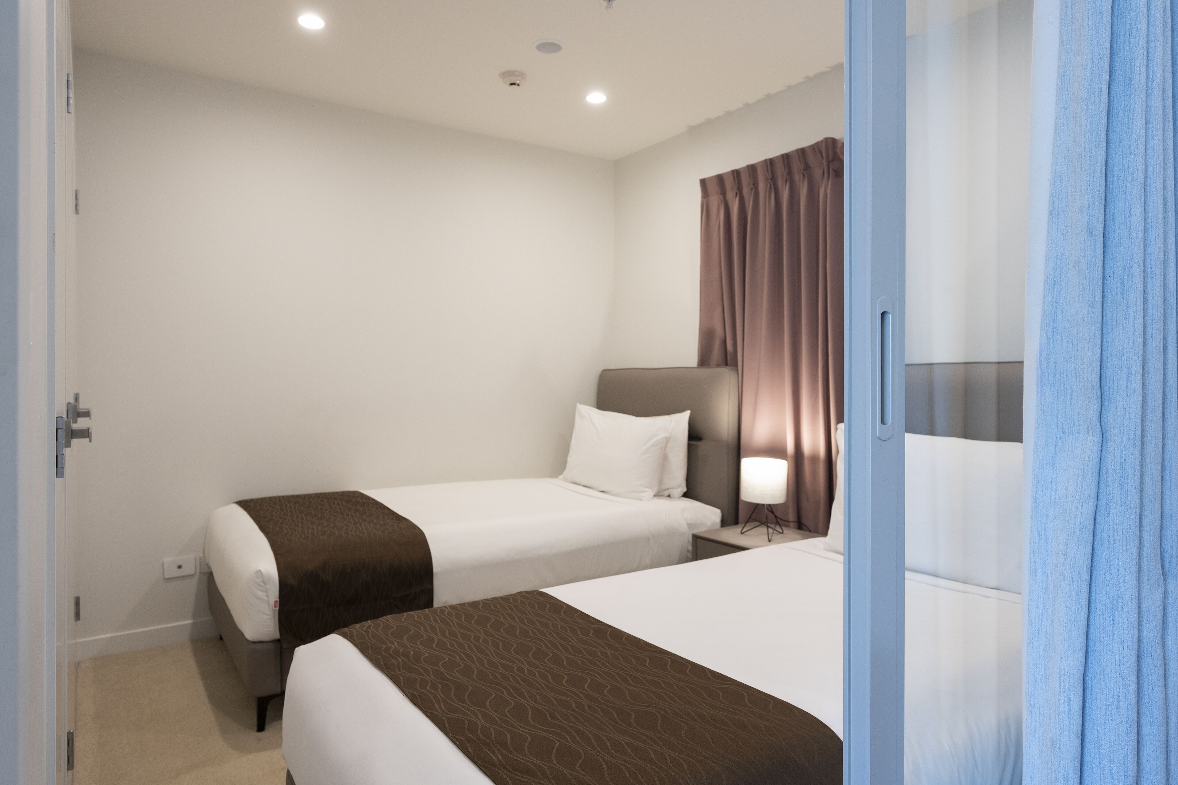 A hotel room with two beds, a nightstand, a lamp, and a window with curtains.