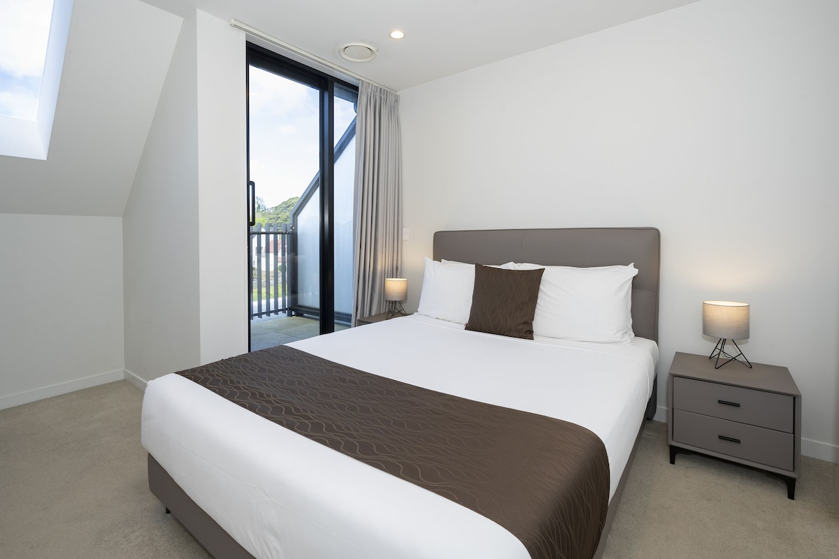 A modern hotel room with a large bed, a nightstand with a lamp, and a sliding glass door leading to a balcony.