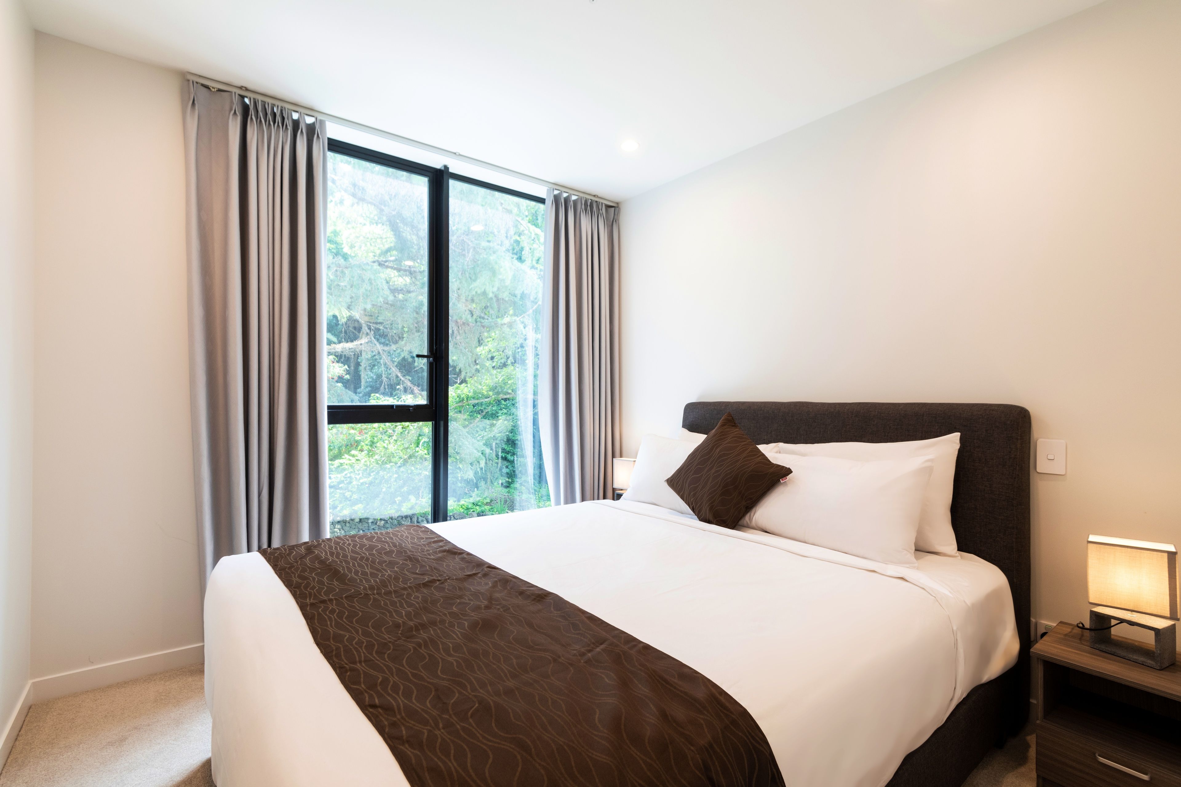 A hotel room with a large bed, a nightstand with a lamp, and a view of greenery through the window.