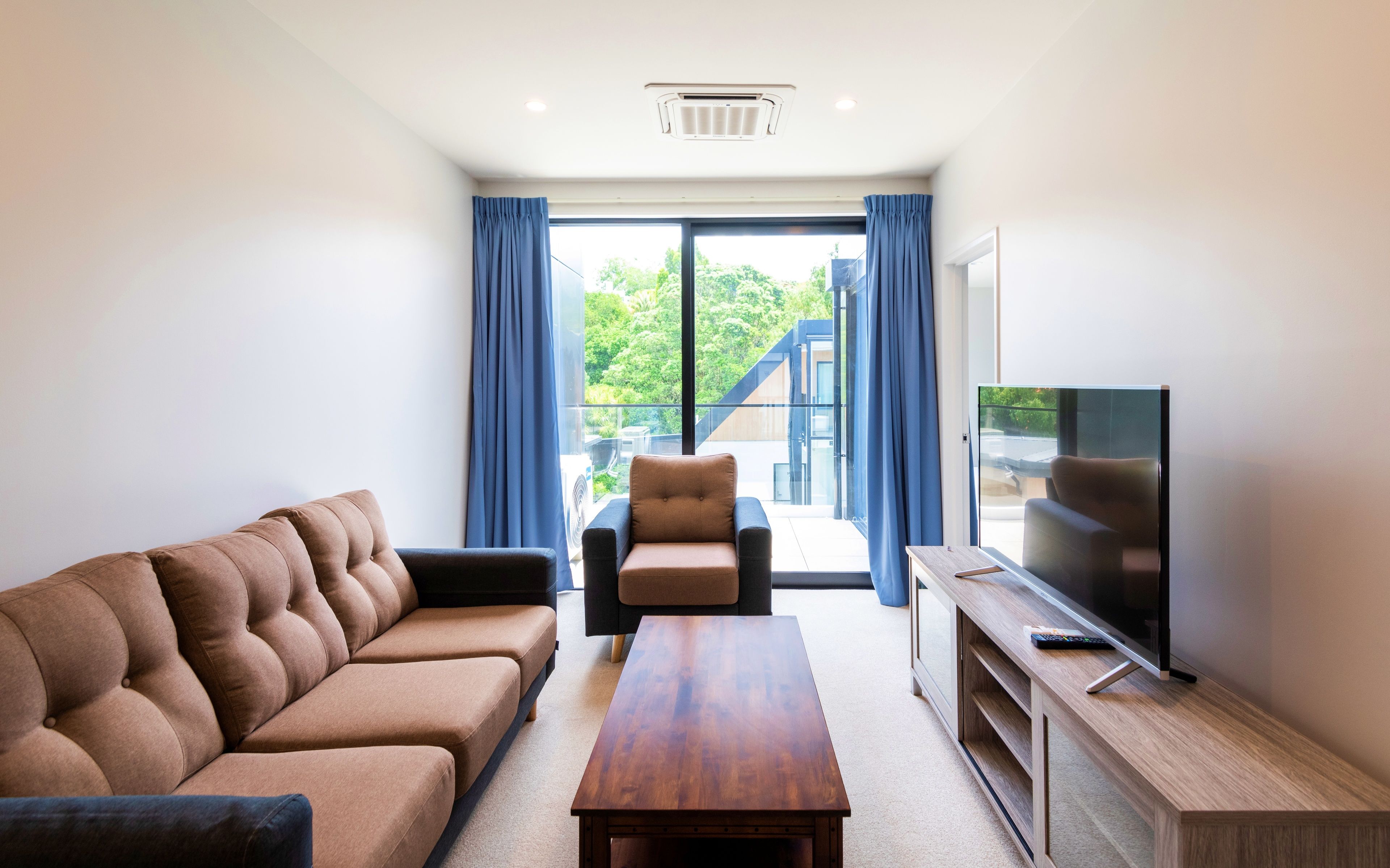 Un soggiorno moderno con un divano marrone, un tavolino in legno, una televisione su un mobile in legno e vista su un balcone con tende blu.