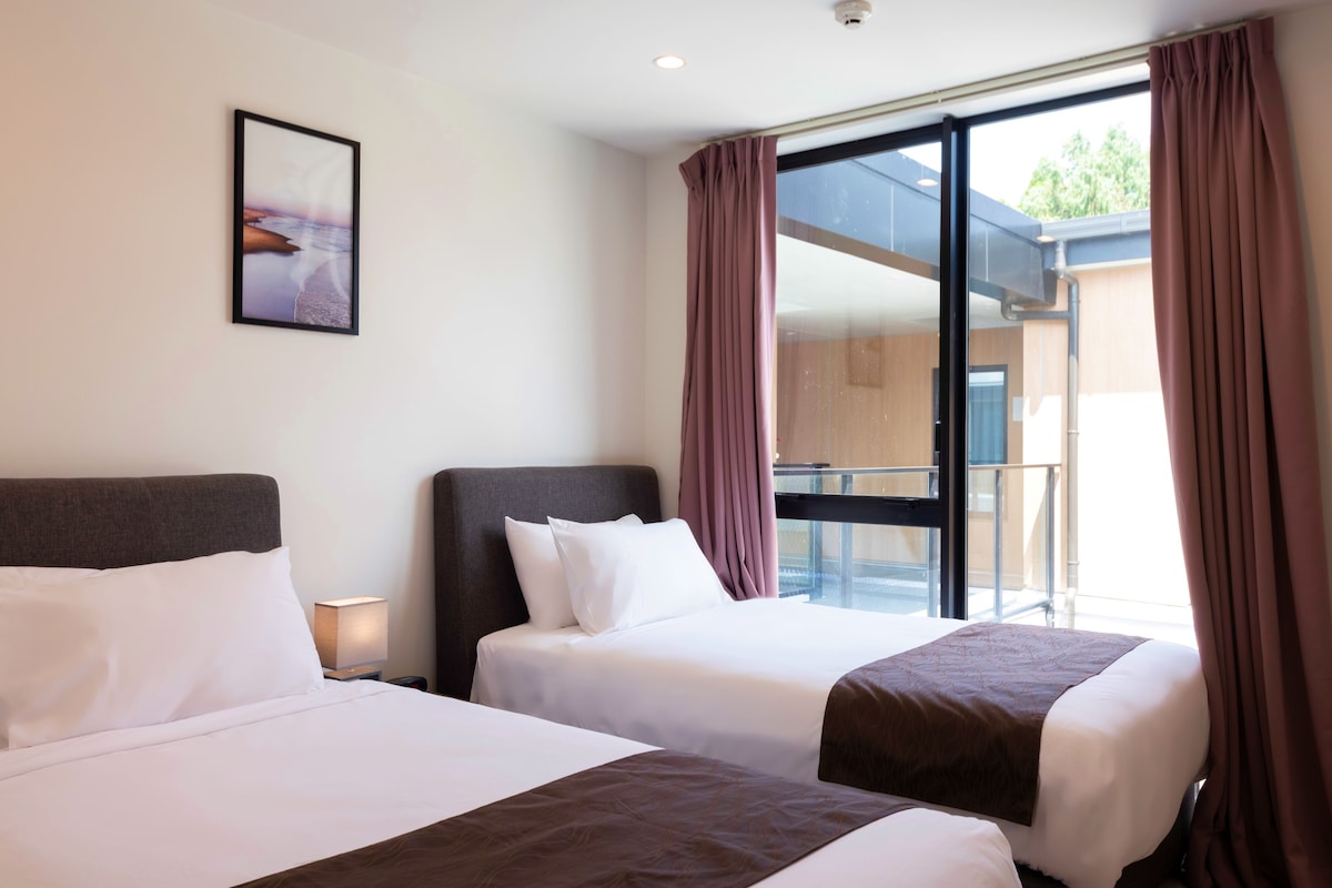 A hotel room with two beds, a nightstand, a framed picture on the wall, and a view of the outdoors through a sliding door.