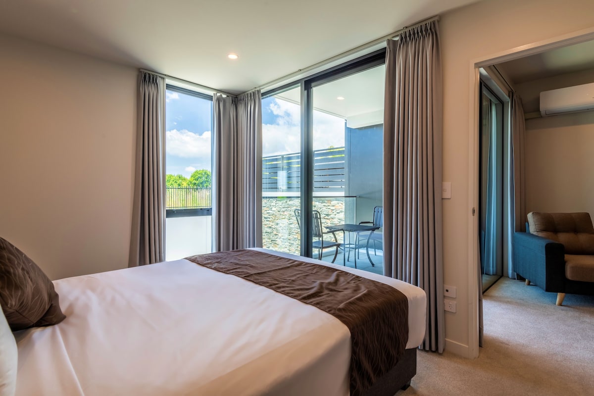 A hotel room with a bed, a balcony with a table and chairs, and an air conditioning unit.
