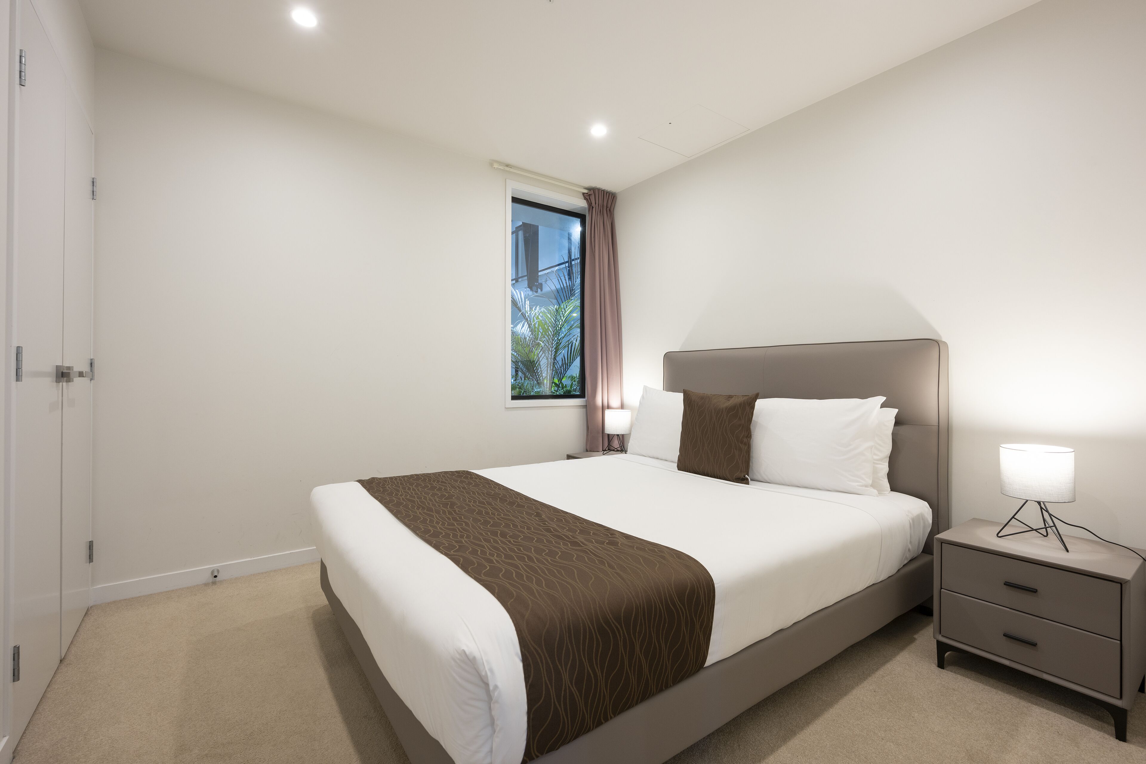 A modern hotel room with a large bed, a bedside table with a lamp, and a window with curtains.