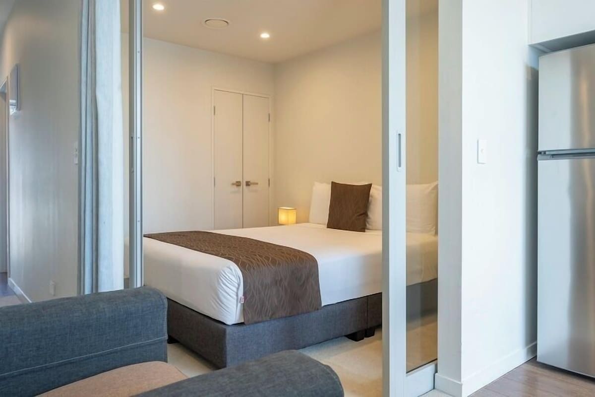 A modern hotel room with a bed, a bedside table, a lamp, and a refrigerator.