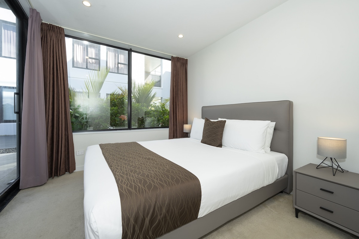 A modern hotel room with a large bed, a bedside table with a lamp, and a view of greenery outside.