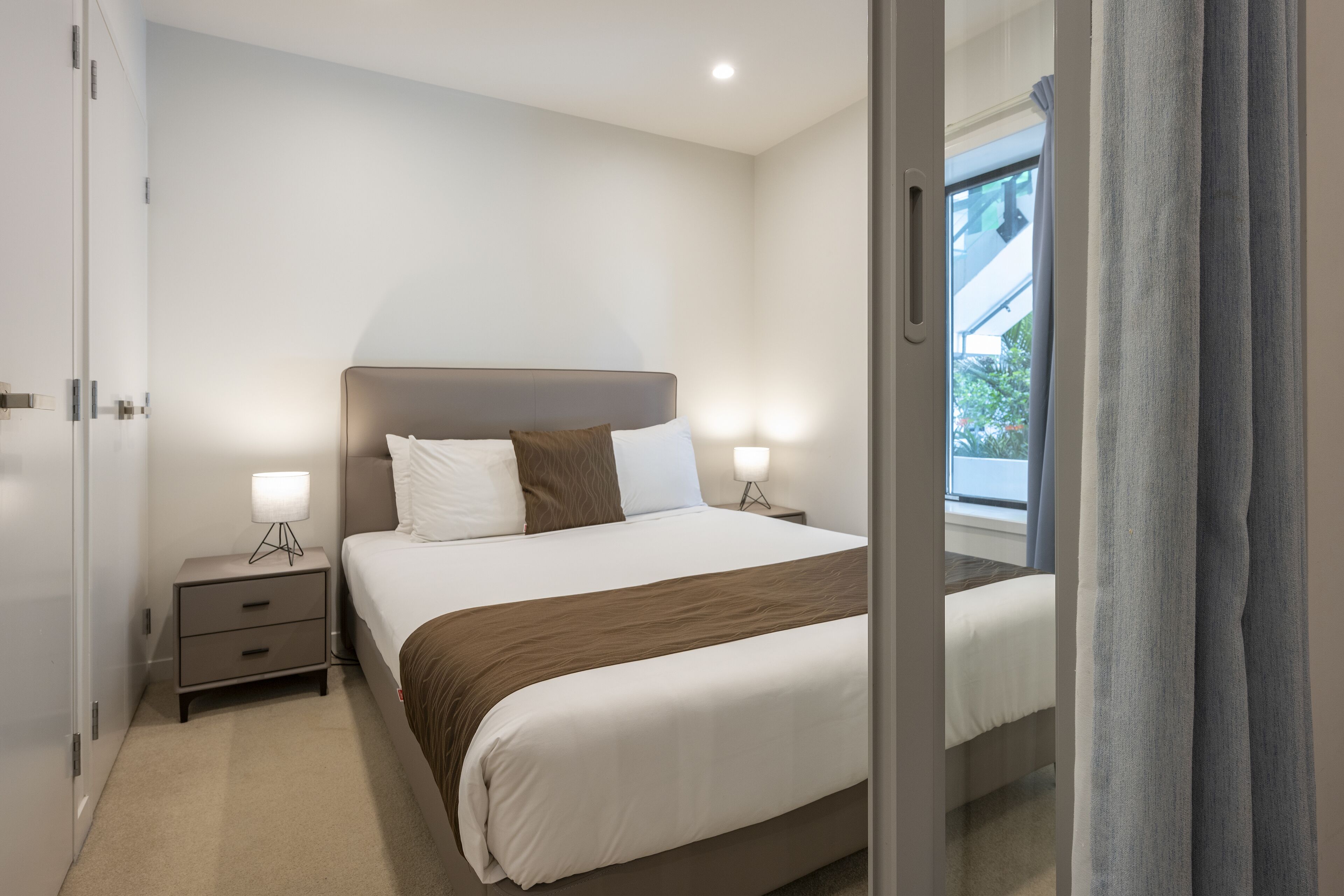 A modern hotel room with a large bed, bedside lamps, a nightstand, and a window with curtains.