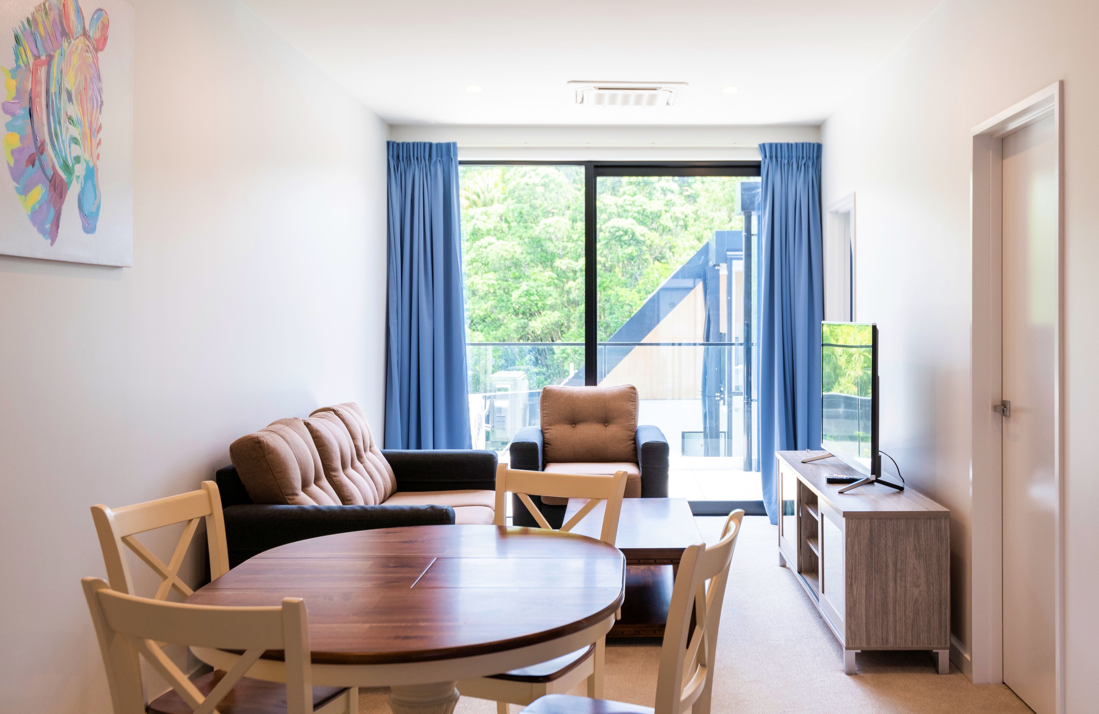 Una sala de estar moderna con una mesa redonda de madera, sillas color beige, un sofá marrón y un televisor sobre un mueble de madera.