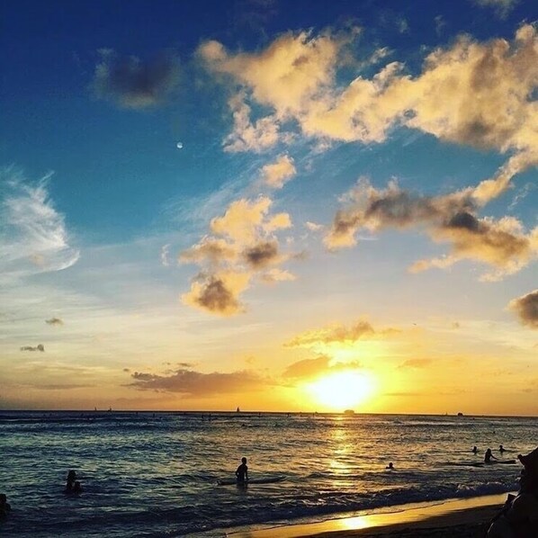 Une scène de plage au coucher du soleil sur l’océan, avec des gens qui se baignent et un ciel dégagé parsemé de nuages.