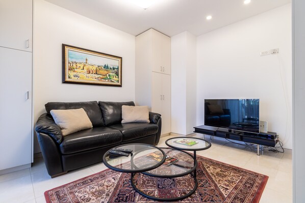 A modern living room with a black leather sofa, two glass coffee tables, a large flat-screen TV, and a patterned rug. - New exclusive 24 Hour Security Building With Shabbat Elevator and large gym (Jerusalem)