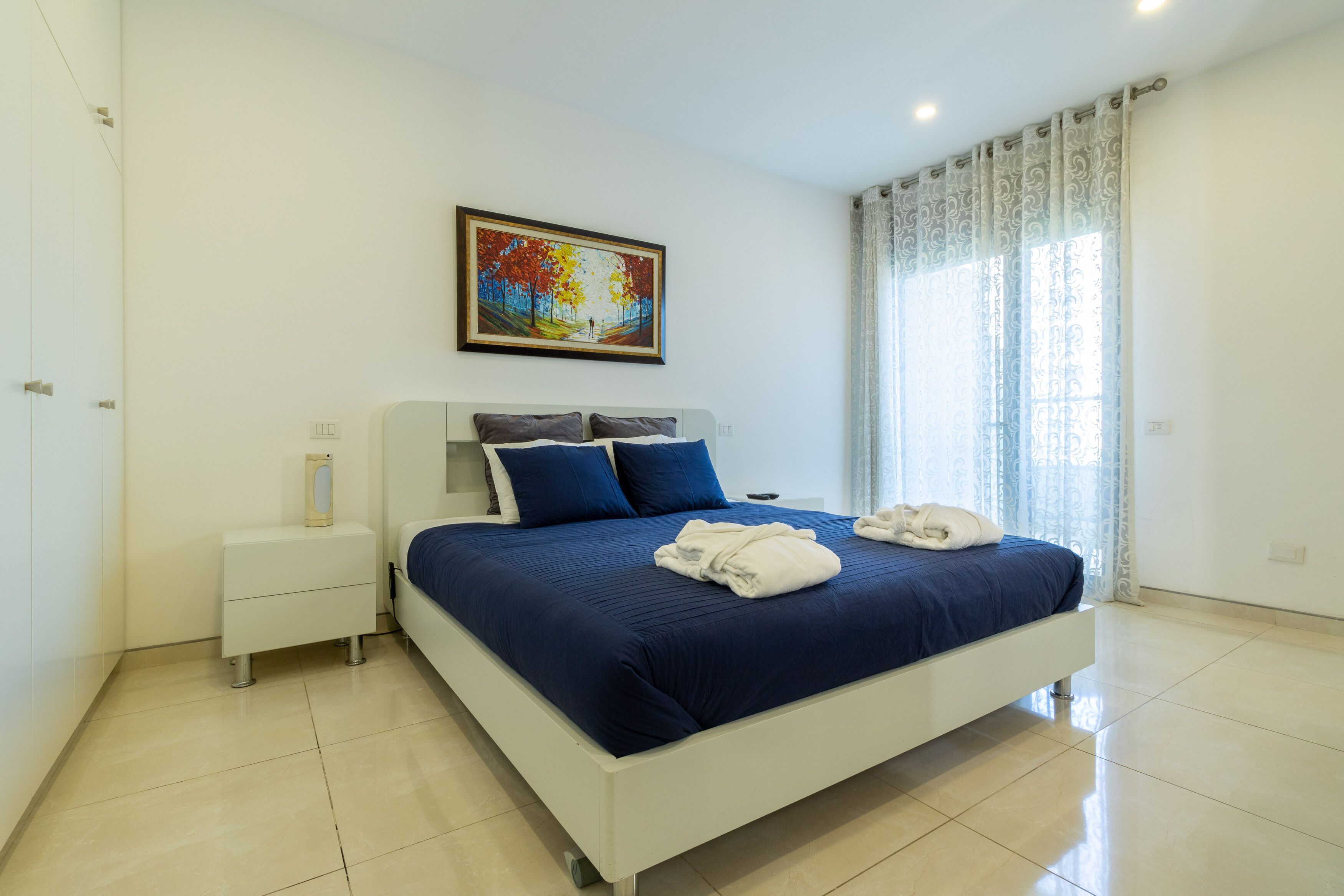 A modern bedroom with a large bed, a nightstand, a bedside table, a framed picture on the wall, and a window with curtains.