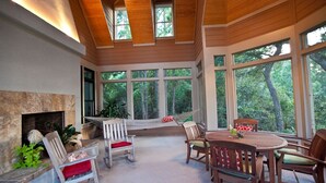 Une vaste terrasse dotée d’une cheminée en pierre, de meubles en bois et de grandes fenêtres offrant une vue sur les arbres environnants.