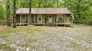 A rustic cabin with a porch, surrounded by trees and a gravel driveway.