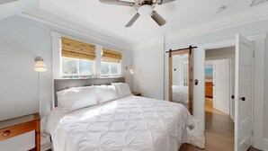 A bedroom with a bed, a ceiling fan, a window with blinds, a bedside lamp, and a door leading to another room.