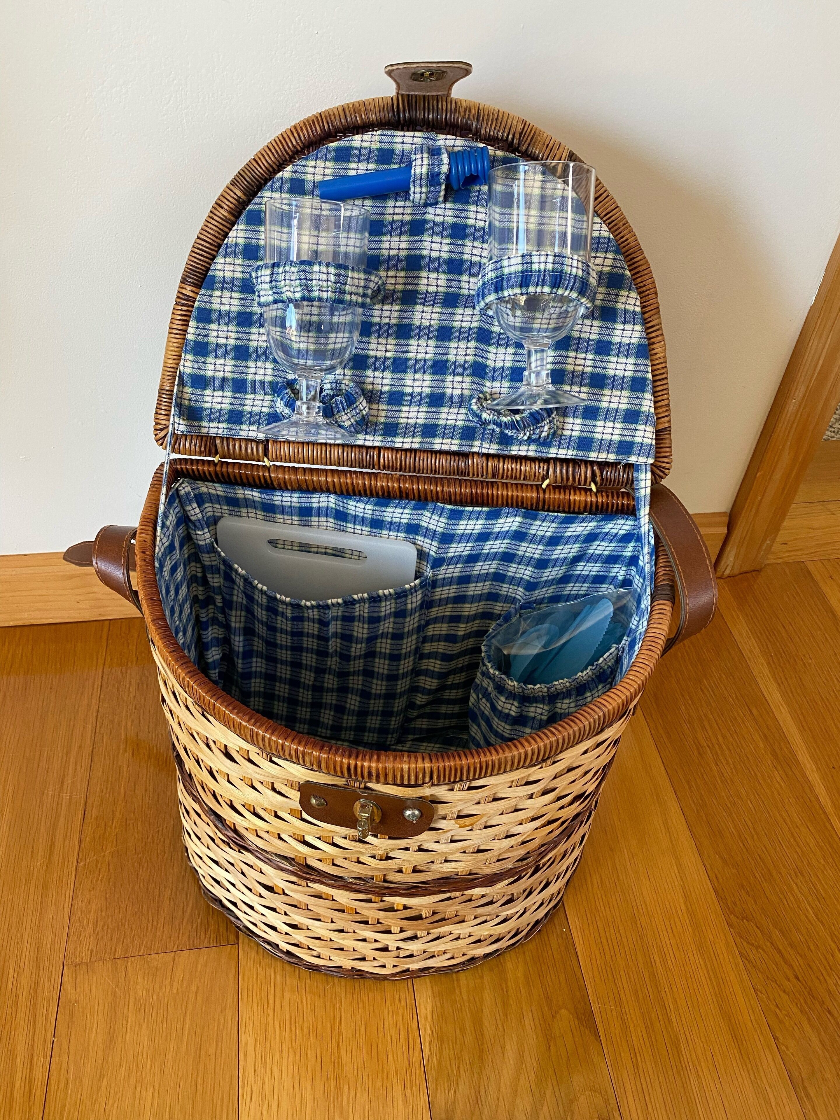 Un cesto di vimini con una coperta a quadri, contenente una bottiglia azzurra, un bicchiere e un sacchetto di plastica.