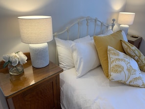 A neatly made bed with white and gold pillows, a wooden nightstand with a lamp, and a vase with white flowers.
