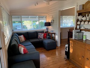 A modern living room with a sectional sofa, a wooden cabinet with kitchenware, and a large window with blinds. - The Old Boathouse - kayak / high speed internet / minutes to the beach (Burrill Lake)