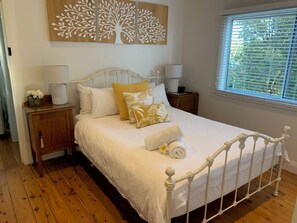 A neatly made bed with white bedding and a white metal frame, a wooden nightstand with a lamp, and a wall-mounted tree decoration.