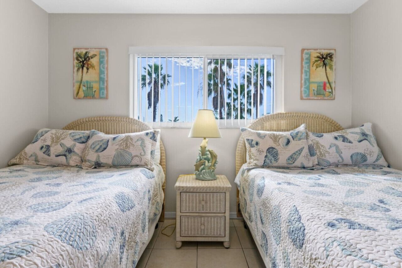 Een slaapkamer met twee bedden, een nachtkastje met lamp en een raam met palmbomen buiten.