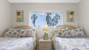 A bedroom with two beds, a nightstand with a lamp, and a window with palm trees outside.