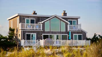 A two-story wooden house with a balcony, large windows, and a view of the beach.