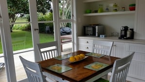 Uma área de refeições com uma mesa de madeira, cadeiras brancas e um banco acolchoado verde. Há também uma cozinha com armários brancos e um micro-ondas.