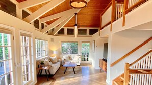 A spacious interior with a high ceiling, wooden beams, a staircase, and a seating area with a table and cushions.