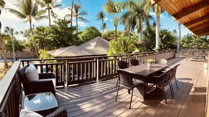 A wooden deck with outdoor furniture, including a table and chairs, surrounded by palm trees and a building in the background.