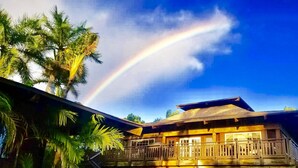 A wooden building with a balcony, surrounded by palm trees, under a clear sky with a rainbow.